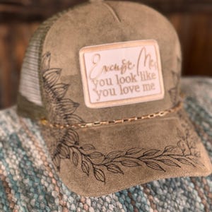 May include: Olive green trucker hat with a white mesh back and a decorative sunflower and leaf design. The hat features a rectangular patch with the text "Excuse Me, You look like you love me."