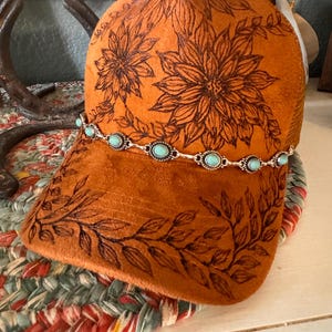 May include: An orange baseball cap with a floral design and a turquoise stone band. The cap features a brown floral pattern on the front and brim. The band is made of silver-colored metal and has oval turquoise stones. The cap has a mesh back.