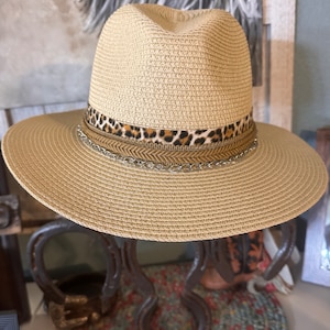 May include: A beige straw hat with a leopard print band and a gold chain accent. The hat is sitting on a stand made of horseshoes.