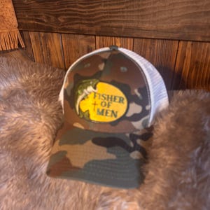 May include: Camouflage baseball cap with a white mesh back. The front of the cap features a yellow oval with the words "FISHER OF MEN" and a fish graphic. The cap is designed for outdoor activities.