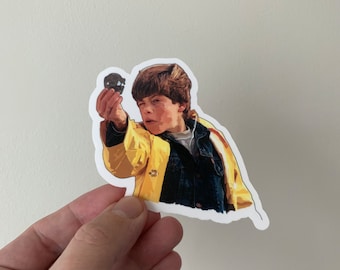 Könnte beinhalten: Ein Aufkleber mit einem jungen Jungen in einem gelben Regenmantel, der ein kleines, rundes Objekt hält. Der Junge schaut aufmerksam auf das Objekt und sein Gesichtsausdruck ist ernst. Der Aufkleber ist auf weißem Papier gedruckt.