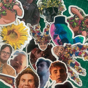 May include: A collection of colorful stickers featuring various characters and designs. The stickers include a sunflower, a character from the movie 'The Shining', a character from the movie 'The Office', and a character from the movie 'The Wolf of Wall Street'.