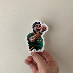 Puede incluir: Una pegatina que representa a un jugador de rugby con una camiseta verde y dorada, celebrando con el puño en alto y la boca abierta. La pegatina tiene un borde blanco y la sostiene una mano sobre un fondo neutro. El jugador lleva un protector de cabeza negro.