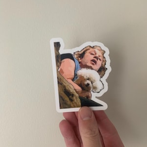 May include: A sticker featuring a cartoon illustration of a man with blonde hair holding a small white dog. The man is wearing a blue shirt and has a surprised expression on his face.