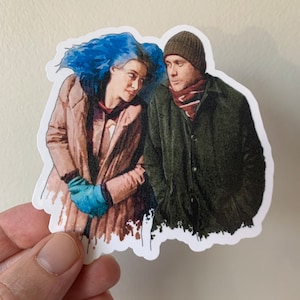 Puede incluir: Una pegatina con una ilustración de acuarela de una pareja. La mujer tiene el pelo azul y lleva un abrigo marrón y guantes azules. El hombre lleva un sombrero marrón, una chaqueta verde y una bufanda roja y blanca.