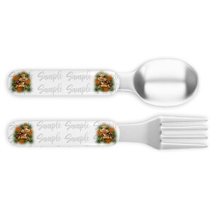 May include: A set of children's cutlery featuring a spoon and fork with a white handle and a colorful cartoon turtle design. The handle has the words "Sample Sample Sample Sample" repeated.