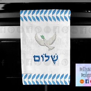 Op de afbeelding: Een witte keukendoek met een blauw-wit patroon. De handdoek heeft een witte duif met een olijftak in zijn bek en het Hebreeuwse woord "Shalom" in blauwe letters. De handdoek hangt aan een zwarte ovendeur.