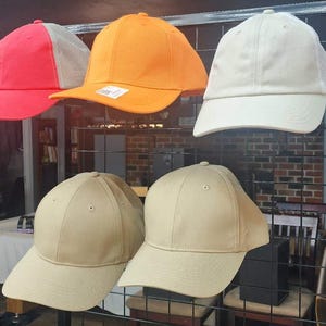 Puede incluir: Exhibición de gorras de béisbol en varios colores, incluyendo rojo, naranja y beige. Algunas gorras tienen la parte trasera de malla. Las gorras se muestran en una rejilla metálica negra.