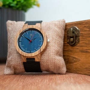 May include: A wooden wristwatch with a blue face and black leather band. The watch is sitting on a brown cushion and comes in a wooden box.