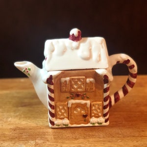 May include: A white ceramic teapot shaped like a gingerbread house with red and white candy cane accents. The teapot has a brown roof with white snow and a red chimney.