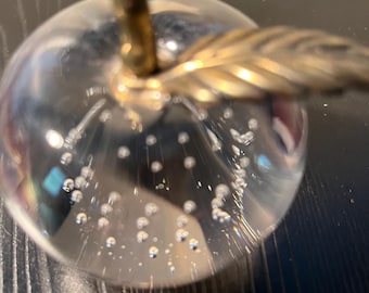 May include: A clear glass apple paperweight with a gold-colored stem and leaf. The apple is filled with small air bubbles, creating a unique visual effect. The paperweight is a decorative item.