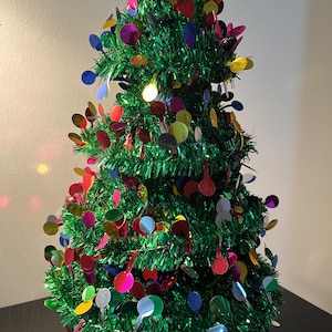 May include: A small artificial Christmas tree decorated with colorful tinsel and round ornaments. The tree is lit with a white light.