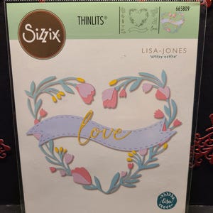 Puede incluir: Un juego de troqueles Sizzix Thinlits con una corona de flores en forma de corazón con la palabra "love" en una pancarta. El juego de troqueles está diseñado por Lisa Jones e incluye 11 troqueles.