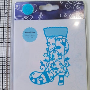 Puede incluir: Una troquelada de metal azul de una bota de duende con un diseño de acebo y bayas. La troquelada mide 6,5 cm por 9,3 cm. El embalaje incluye el texto "Rococo Studios" y "Pixie Boot".