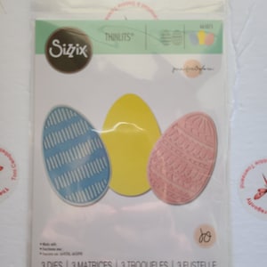 Puede incluir: Un paquete de tres troqueles Sizzix Thinlits para manualidades. Los troqueles tienen forma de huevos de Pascua con diferentes patrones: rayas azules y blancas, amarillo sólido y rosa con un diseño floral. El paquete incluye el texto "3 DIES | 3 MATRICES | 3 TROQUELES | 3 FUSTELLE | 3 ВЫРУБКИ | 3 STANZEN | 3 CORTANTES".