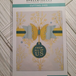 May include: A Spellbinders paper arts cutting die set featuring a butterfly with a bow, flowers, and a tag that says "From All Of Us". The die set is designed to create a card or scrapbook layout.