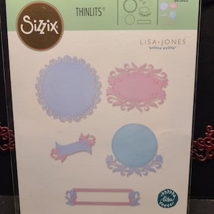 Puede incluir: Un paquete de troqueles Thinlits de Sizzix de Lisa Jones. El paquete contiene ocho troqueles de varias formas, incluyendo círculos, óvalos y una pancarta. Los troqueles son en tonos azules y rosas. El paquete también incluye el texto "8 DIES | 8 MATRICES | 8 TROQUELES | 8 FUSTELLE".