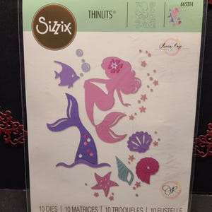 Op de afbeelding: Een pakket met 10 uitgestanste metalen sjablonen voor knutselen. De sjablonen tonen een zeemeermin met een staart, schelpen, zeesterren en bubbels. De verpakking is gelabeld met "Sizzix Thinlits" en "Olivia Rose".