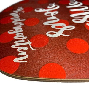 May include: Red apple-shaped wooden sign with white polka dots and the text "Mrs. Jones' Kindergarten" in white lettering.