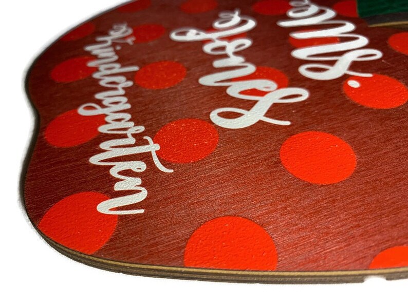 May include: Red apple-shaped wooden sign with white polka dots and the text "Mrs. Jones' Kindergarten" in white lettering.