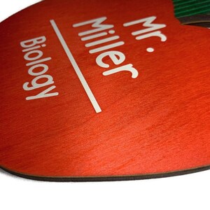 May include: Red apple-shaped wooden sign with white text that reads "Mr. Miller Biology".