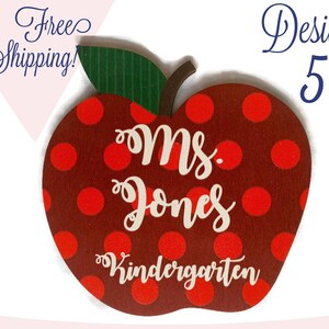 May include: Red apple-shaped wooden sign with white polka dots. The sign says "Ms. Jones Kindergarten" in white lettering.