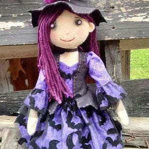 Puede incluir: Una muñeca de bruja morada y negra con un sombrero de bruja negro, un vestido morado con diseños de murciélagos y medias rayadas blancas y negras.