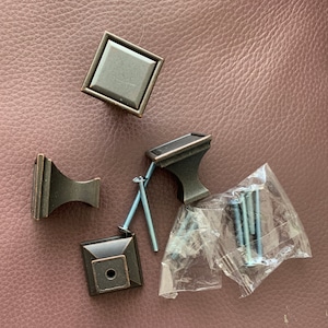 May include: Four bronze cabinet knobs with a square or rectangular shape and a single screw hole. The knobs are arranged on a brown textured surface with a clear plastic bag containing screws.