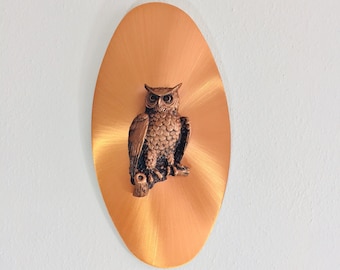 May include: A copper-coloured wall hanging with an owl sculpture. The owl is perched on a branch, set against an oval-shaped background with a radial brushed pattern. The owl is brown with detailed feathers.