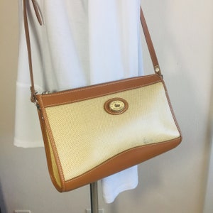 May include: A tan and brown crossbody bag with a woven fabric body and brown leather trim. The bag has a gold metal logo with the text "Dooney & Bourke" on it.