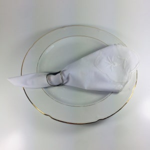 May include: A white napkin with an embroidered design is placed on a white plate with a gold rim.