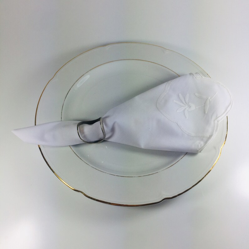 May include: A white napkin with an embroidered design is placed on a white plate with a gold rim.