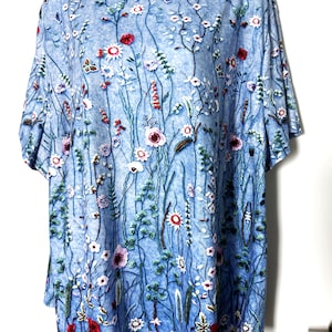 May include: A light blue, short-sleeved top with a floral pattern. The design features a variety of flowers in red, pink, and white, with green stems and leaves. The top has a round neckline and a loose fit.