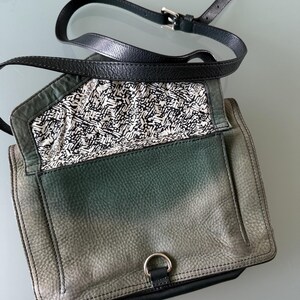 May include: A green and black leather crossbody bag with a fold-over flap, a silver-toned metal D-ring closure, and an adjustable black leather strap. The interior lining features a repeating black and white graphic pattern with the text REBECCAMINKOFF.