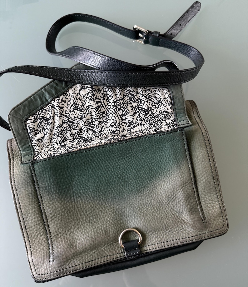May include: A green and black leather crossbody bag with a fold-over flap, a silver-toned metal D-ring closure, and an adjustable black leather strap. The interior lining features a repeating black and white graphic pattern with the text REBECCAMINKOFF.
