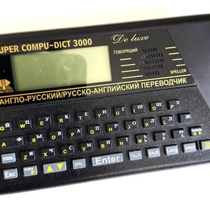 May include: Black Super Compu-Dict 3000 electronic translator with a small screen and a keyboard featuring both English and Cyrillic characters. The device has the text "De Luxe" and "SPELLER" printed on it.