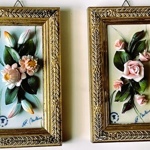May include: Two framed ceramic floral wall plaques with gold frames. The plaques feature 3D pink flowers and green leaves on a white background. The plaques are signed "R. Martini".