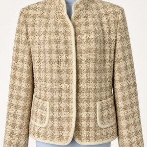 May include: A beige and tan tweed blazer featuring a textured plaid pattern, a stand-up collar, and two front patch pockets with cream-colored trim. The jacket has long sleeves and a straight hemline, finished with matching cream piping along the edges.
