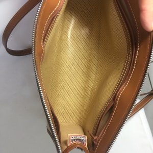 May include: Inside view of a brown leather handbag with a zipper closure. The interior is beige with a textured fabric lining. A small brown leather tag with the text "DOONEY & BOURKE Made in USA" is visible.