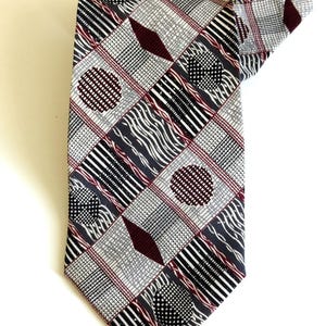 May include: A patterned necktie featuring a geometric design with squares, circles, and diamond shapes. The tie's color palette includes gray, black, white, and burgundy. The tie is made of a smooth fabric.
