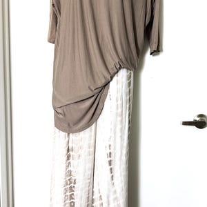 May include: A taupe short-sleeved top with a round neckline and a draped, asymmetrical hem paired with wide-leg trousers featuring a white and taupe vertical tie-dye pattern. The ensemble is displayed on a mannequin against a white door.