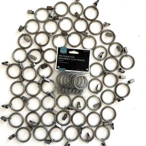 May include: A collection of pewter-colored decorative drapery clip rings. The rings are arranged in a circular pattern on a white surface. A package of seven rings is visible, with the text "Decorative Drapery Clip Rings". Each ring has a clip attached.