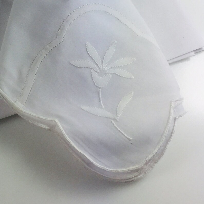 May include: A stack of white cotton napkins with a delicate embroidered floral design. The napkins have scalloped edges and are perfect for adding a touch of elegance to any table setting.