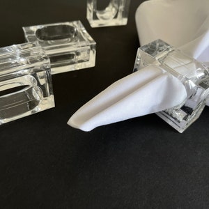 May include: Four clear acrylic napkin rings, one with a white napkin placed through it. The napkin rings are square shaped with a rounded opening for the napkin.