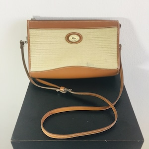May include: A brown leather crossbody bag with a beige canvas front panel. The bag has a Dooney & Bourke logo on the front. The bag has a long brown leather strap.