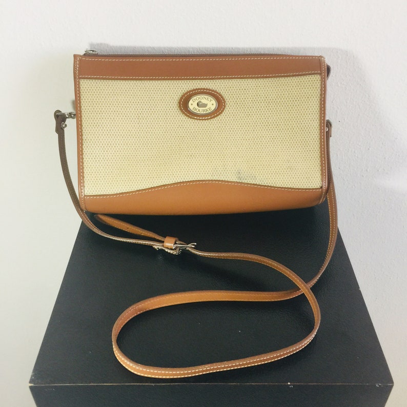 May include: A brown leather crossbody bag with a beige canvas front panel. The bag has a Dooney & Bourke logo on the front. The bag has a long brown leather strap.