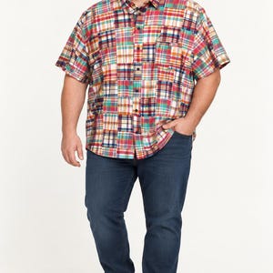 May include: A short-sleeved button-down shirt featuring a multi-colored patchwork plaid pattern in shades of red, blue, yellow, and white. The shirt is paired with dark blue denim jeans and plain white sneakers, creating a casual ensemble.