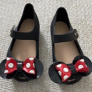 May include: Black jelly shoes with a red and white polka dot bow. The shoes have a strap across the top and a small buckle.