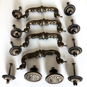 May include: A collection of antique bronze-colored cabinet hardware, including four ornate drawer pulls and several matching knobs. The pulls have a curved design with decorative detailing, and the knobs feature a floral pattern.