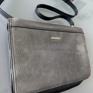 May include: A rectangular crossbody bag featuring a textured grey leather flap and a smooth black leather base. The bag includes a thin black adjustable shoulder strap with a silver-toned metal buckle and a small rectangular silver logo plate.